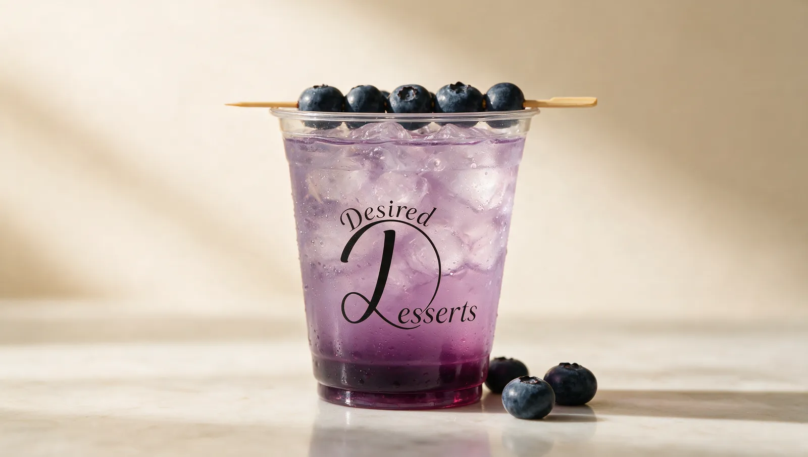 Desired Desserts Berry Bliss signature sip — lilac blueberry lemonade over ice in a clear branded cup, topped with a skewer of fresh blueberries.